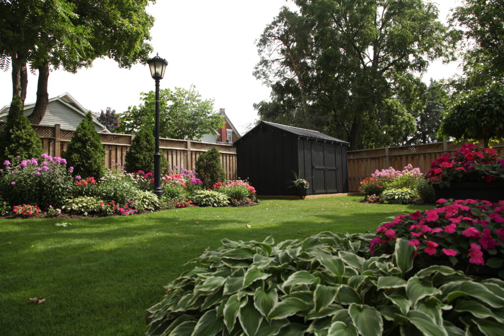 Dark grey storage shed installed in a landscaped backyard with green lawn and wood-fence backdrop